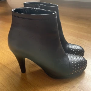 Svarta stövletter med nitar och klack - Snygga svarta stövletter i skinn med hög klack och rund tå. Tåpartiet är dekorerat med silvriga nitar som ger en cool edge. Dragkedja på insidan för enkel på- och avtagning. Perfekta för dig som vill sticka ut med detaljer. Endast provade, aldrig använda. 