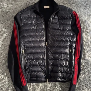 Svart Moncler pufferjacka med röda detaljer - Size M. Nypris ligger runt 12000kr, passa på! Svart pufferjacka från Moncler med glansig, quiltad front och ärmar i mjuk bomull. Röda ränder och Moncler-logga längs ärmarna ger en sportig vibe. Jackan har dragkedja framtill och två fickor med dragkedja. Perfekt för dig som gillar streetstyle.