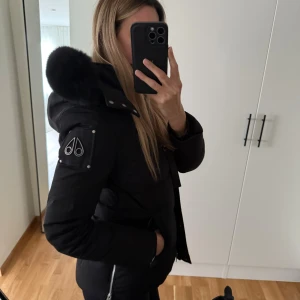 Svart dunjacka Moose Knuckles L - Svart dunjacka från Moose Knuckles i storlek L med fluffig päls på huvan och snygga metalldetaljer på ärmen. Jackan har dragkedja, långa ärmar och en stilren, quiltad design som håller dig varm hela vintern. Den är använd en vinter, finns lite tecken på användning på ärmarna (för bilder hör av dig). Köpt på Johnells, upplevs mindre i storlek snarare mer som en M 