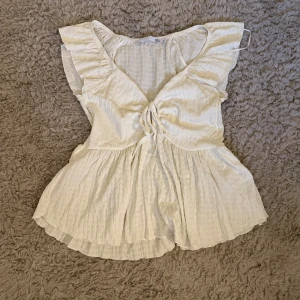 Vit blus med volang och snörning - Supersöt vit blus från 157 med volangdetaljer vid axlarna och v-ringning med snörning framtill. Blusen har en luftig, lätt rynkad struktur och är perfekt för varma dagar. Den är ärmlös och har en flowy passform som ger en romantisk vibe.