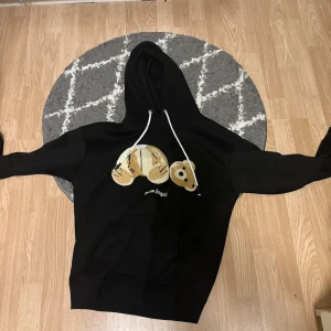 Palm Angels svart hoodie med björn - Svart hoodie från Palm Angels med ikoniskt björnmotiv framtill. Tröjan har huva med vita snören och klassisk känguruficka. Perfekt för dig som gillar streetwear och vill sticka ut med en cool detalj på bröstet.