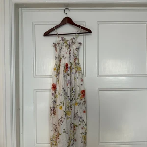 Blommig långklänning med smock - Superfin långklänning med smala axelband och smock upptill. Klänningen är vit med färgglatt blommigt mönster i rött, gult, lila och grönt. Den har en luftig och flowy passform, perfekt för varma dagar. Materialet känns lätt och somrigt.