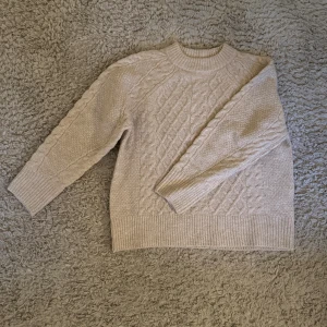 Beige stickad tröja från 157 - Mysig beige stickad tröja från 157 med klassiskt flätmönster och ribbade muddar. Tröjan har rund halsringning och långärmade ärmar, perfekt för lager på lager under kyliga dagar.