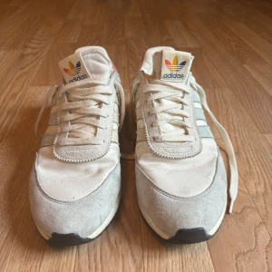 Adidas I-5923 sneakers | Love Unites - Säljer ett par Adidas I-5923 sneakers med retrovibbar. Skorna är vita med ljusgrå och rosa detaljer, har klassiska tre ränder på sidorna och Adidas-logga på plös och häl. Ovandel i textil och mocka, mellansula i boost-material för extra komfort. Perfekta för dig som gillar old school-stil.