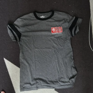 Grå Hollister t-shirt med svart kant - Snygg grå t-shirt från Hollister med svart kant vid halsen och ärmsluten. Tryck med röd och vit logga på bröstet. Klassisk passform och mjukt bomullsmaterial, perfekt för en chill stil.