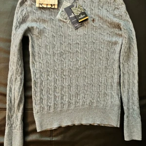 Blå Morris tröja kabelstickad - Supersnygg kabelstickad tröja från Morris i ljusblå merinoull. Klassisk rund halsringning och diskret broderad logga på bröstet. Perfekt för dig som gillar stilrena och tidlösa plagg med premiumkänsla. Långärmad och riktigt mjuk mot huden.