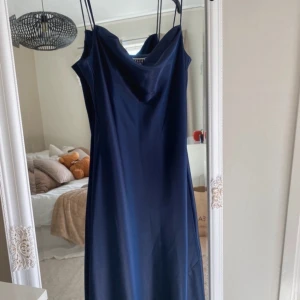 Mörkblå balklänning från Yessica - Elegant mörkblå balklänning med smala axelband och draperad halsringning. Perfekt för en stilren look. Klänningen har en mjuk och glansig yta som ger en lyxig känsla.