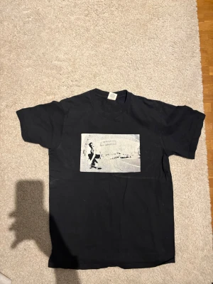 Supreme svart t-shirt med skate-tryck - Svart t-shirt från Supreme i 100% bomull med ett stort svartvitt tryck av en skateboardåkare på bröstet. Klassisk passform med korta ärmar och rund hals. Liten Supreme-logga i nacken. Perfekt för dig som gillar streetwear och skatekultur.