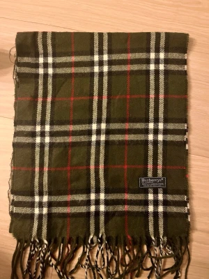 Grön rutig Burberrys väst i ull - Snygg väst från Burberrys i klassiskt rutigt mönster med gröna, vita, svarta och röda toner. Tillverkad i 100% lammull, mjuk och varm. Västen har fransar nedtill och en diskret Burberrys-label. Perfekt för dig som gillar tidlös stil och vill sticka ut.