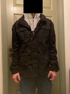 Svart fieldjack från Brandit - Säljer en riktigt fet svart fieldjacka från Brandit med fyra stora fickor framtill, axelklaffar och dragkedja samt knappar framtill. Jackan har en snygg krage med inbyggd huvva bakom en dragkedja. Vid funderingar, fråga på! 