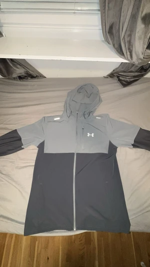 Grå och svart vindjacka Under Armour - Snygg vindjacka från Under Armour i storlek L. Jackan är tvåfärgad med grått upptill och svart nedtill, har huva och hel dragkedja framtill.  jackan har en sportig look med logga på bröstet och praktiska fickor.