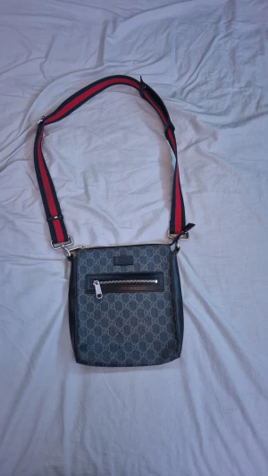 Svart axelväska - Säljer en snygg axelväska. Med bred axelrem i rött och marinblått, samt detaljer i svart skinn. Framsidan har en praktisk dragkedja och insidan är fodrad med svart tyg. Perfekt för dig som gillar designer och vill sticka ut.