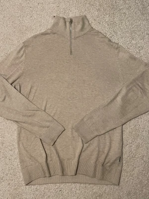 Jack & Jones Zip Tröja - Säljer en beige långärmad tröja från Jack & Jones med half zip i halsen. Tröjan har ribbade muddar vid ärmslut och nederkant, samt en hög krage. Materialet är mjuk bomull och passformen är normal. Perfekt för dig som gillar stilrena och enkla plagg.