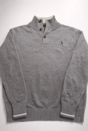 Grå stickad tröja från Polo Ralph Lauren - Snygg grå stickad tröja från Polo Ralph Lauren med ribbad hög krage och tre knappar framtill. Tröjan har långärmade muddar med vit rand och klassisk Polo-logga broderad på bröstet. Perfekt för kyliga dagar och stilrena outfits.