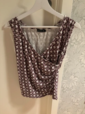 Mönstrad omlottopp från Stockh LM - Säljer en ärmlös omlottopp från Stockh LM i storlek S. Toppen har ett geometriskt mönster i vinrött och vitt, med v-ringad front och draperad effekt. Perfekt för dig som gillar unika detaljer och snygg passform.