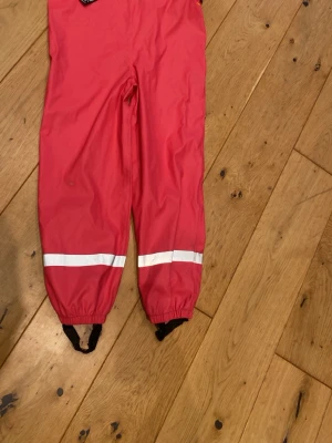 Rosa regnbyxor från 157 Funk - Säljer ett par rosa regnbyxor från 157 Funk med svarta hängslen med logga, elastiska benslut och reflexband runt benen. Perfekta för regniga dagar och har justerbara hängslen för extra komfort. Materialet är slitstark polyester.