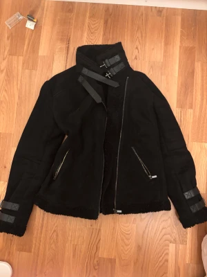 Svart shearlingjacka - Svart shearlingjacka med krage och mjukt foder. Jackan har två dragkedjefickor, snygga spännen vid halsen och på ärmarna samt råa kanter. Perfekt för dig som gillar en cool och mysig stil med extra detaljer.