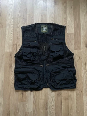 Väst - Säljer en svart outdoor-väst i storlek M med flera praktiska fickor och dragkedjor framtill. Västen har meshpartier för extra ventilation och är perfekt för dig som gillar friluftsliv eller fiske.