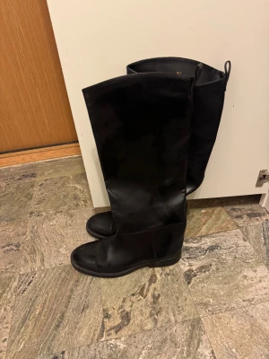 Svarta höga skinnstövlar (ej äkta skinn) - Säljer ett par svarta stövlar i skinn (ej äkta skinn)  med rund tå och platt sula. Stövlarna är höga och har en stilren, minimalistisk design utan synliga detaljer. Perfekta för dig som gillar klassiska boots med enkel look. Kostar 800kr nya men är slut på hemsidan