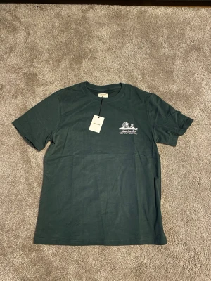 Mörkgrön Aimé Leon Dore t-shirt - Snygg mörkgrön t-shirt från Aimé Leon Dore med vit tryckt grafik och logga både fram och bak. Klassisk rund hals och korta ärmar. Materialet är mjuk bomull som känns skönt mot huden. Perfekt för dig som gillar streetwear och vill ha något stilrent med cool branding.