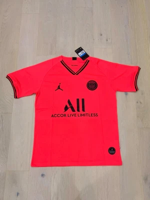 PSG x Jordan röd fotbollströja M - Snygg röd PSG x Jordan fotbollströja i medium med svarta detaljer och loggor. Tröjan har V-ringad hals och korta ärmar med svarta ränder vid mudden. Materialet är lätt och andas, perfekt för fotboll eller träning. Officiell produkt med Paris Saint-Germain emblem och Jordan Jumpman.