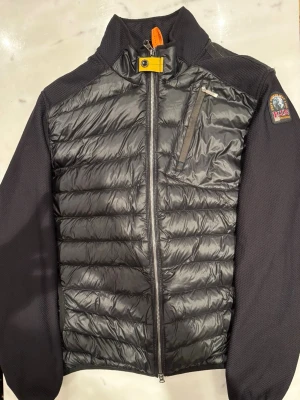 Parajumpers cardigan  - Svart pufferjacka från Parajumpers med quiltad framsida och rygg, samt släta svarta ärmar. Jackan har dragkedja framtill, bröstficka med dragkedja och parajumpers klassiska gula detalj vid halsen. Perfekt för dig som vill ha en snygg och funktionell jacka.