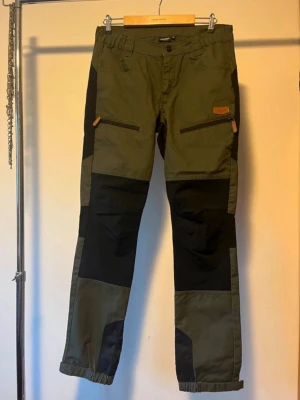 Kayoba outdoor cargopants i grön och svart - Säljer ett par Kayoba outdoor cargopants i grön och svart med förstärkta partier och dragkedjefickor framtill. Byxorna har justerbara benslut och är tillverkade i slitstark bomull och polyester, perfekta för utomhusaktiviteter. Snygg och funktionell design.