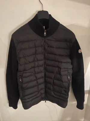 Svart Moncler Cardigan - Svart pufferjacka från Moncler med quiltad framsida och släta ärmar. Jackan har dragkedja framtill, två sidofickor med dragkedja och Moncler-logga på vänster ärm. Materialet är en mix av polyester och ull, perfekt för kyliga dagar.
