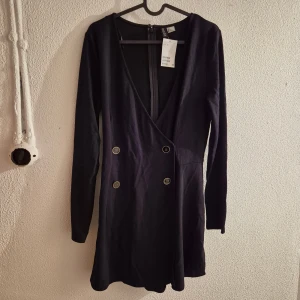 Svart omlottklänning från H&M - Snygg svart kort omlottklänning från H&M med lång ärm och dekorativa knappar framtill. Klänningen har en djup v-ringning och dragkedja i ryggen. Perfekt för dig som gillar stilrena och moderna plagg med en twist..  Nytt med lappen kvar. Jag kan inte se några hål eller skador etc. Även om de är nytt.  Obs! Jag står inte för frakbolagets slarv, skador eller annat som händer efter att jag har postat.