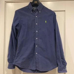 Blå skjorta från Polo Ralph Lauren - Snygg marinblå skjorta från Polo Ralph Lauren i slim fit. Klassisk button-down krage, vita knappar och långärmad design. Broderad grön logga på bröstet. Tillverkad i mjuk bomull som känns skön mot huden.