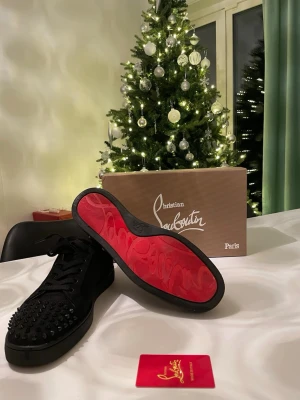 Christian Louboutin svarta sneakers med nitar - Säljer ett par svarta sneakers från Christian Louboutin med ikonisk röd sula och svarta nitar på tån. Skorna har snörning och är tillverkade i mocka med platt sula och rund tå. Allt original medföljer: box, certifikat, extra nitar och kvitto.