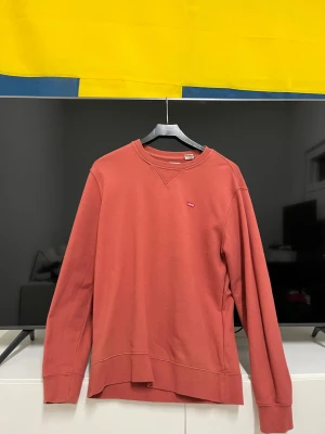 Röd sweatshirt från Levi's - Säljer en röd sweatshirt från Levi's med klassisk crewneck och liten logotyp på bröstet. Tröjan har lång ärm och ribbade muddar vid ärmslut och nederkant. Perfekt för dig som gillar enkel och stilren streetwear.