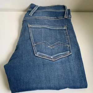 Blå Replay anbass jeans - Säljer ett par blå jeans från Replay med snygga slitningar och ljusare partier på låren. Klassisk femficksmodell med dragkedja och knapp. Jeansen har en smal passform och är tillverkade i mjukt denimtyg som sitter skönt hela dagen. Säljer de billigt eftersom att de är ganska slitna.