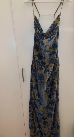 PrettyLittleThing lång klänning blå/guld | satinlook | EU 38 - Elegant långklänning från PrettyLittleThing i blått & guld med blommigt mönster. Smickrande draperad ringning, tunna justerbara axelband och lätt satinliknande material som faller fint. Perfekt till fest, bröllop, sommarmiddag eller semester. Storlek EU 38 (M). Fint skick! ✨