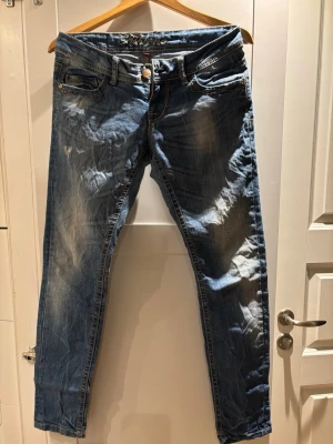 Blå jeans från Fishbone, storlek 26 - Snygga blå jeans från Fishbone i storlek 26. Klassisk femficksmodell med slitningar och kontrastsömmar. Jeansen har låg midja och raka ben, tillverkade i slitstarkt denimtyg. Perfekt för dig som gillar en avslappnad och cool stil.