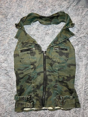 Camouflagetopp med dragkedja och bälten - Säljer en grön camouflagetopp med coola detaljer som dubbla fickor med dragkedja på bröstet, bred krage med kristaller och justerbara bälten. Toppen har en hel dragkedja framtill och är gjord i ett slitstarkt bomullsmaterial. Perfekt för dig som gillar streetstyle. 95% bomull och 5% spandex 