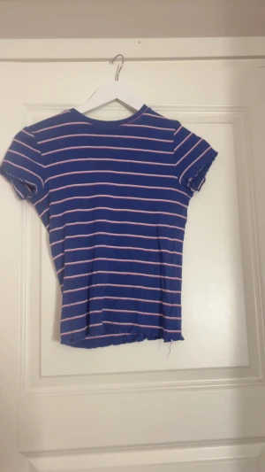 Blå randig croppad topp - Säljer en blå croppad topp med rosa och vita ränder. Toppen har korta ärmar med lätt volangkant och rund halsringning. Materialet känns mjukt och stretchigt, perfekt för en chill look. Passar dig som gillar färg och mönster.
