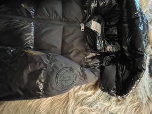 Svart Moncler pufferjacka M - Svart pufferjacka från Moncler i glansigt material med huva och dragkedja framtill. Jackan har Moncler-logga på ärmen och quiltad design som ger en cool look. Perfekt för kalla dagar när du vill vara både varm och snygg.
