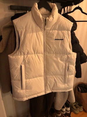 Vit dunväst från Pegador, storlek L - Säljer en stilren vit dunväst från Pegador i storlek L. Västen har hög krage, två sidofickor med dragkedja och ett quiltat mönster. Materialet är polyester och den har en snygg Pegador-logga på bröstet. Perfekt för lager-på-lager och streetwear. Använd en gång!
