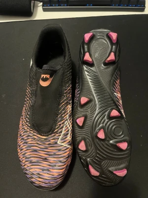 Svarta och lila fotbollsskor från Puma - Säljer ett par fotbollsskor från Puma med svart bas och lila samt orange vågigt mönster. Skorna har en strömlinjeformad design utan snörning och rosa dobbar på sulan. Perfekt för dig som vill sticka ut på planen med snygg stil och bra grepp.