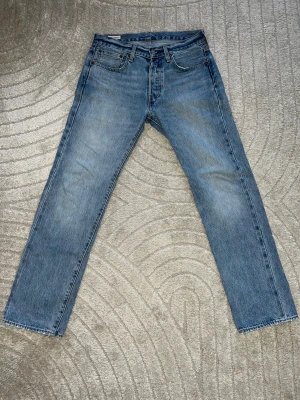 Levi's 501 blå jeans straight fit - Klassiska Levi's 501 jeans i ljusblå denim med raka ben och femficksmodell. Jeansen har knappgylf, läderpatch bak och slitna detaljer vid benslut. Perfekt för dig som gillar en tidlös och avslappnad look.