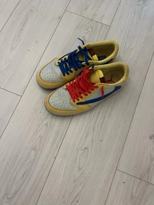 Nike Jordan 1 Travis Scott gul/blå - Nike Jordan 1 Travis Scott sneakers i en unik färgkombo med gul och blå paneler, vit tåbox och kontrasterande röda och blå skosnören. Låg modell med rund tå och platt sula. Insidan har Travis Scott-tryck. Perfekt för dig som gillar färgstarka och hypade sneakers.