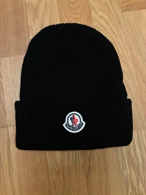 Svart Moncler mössa - Bilderna visar en svart stickad mössa från Moncler med uppvikt kant och logotyp framtill. Passar perfekt som julklapp