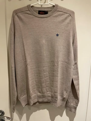 Merino tröja från Morris i storlek M - Snygg beige stickad tröja från Morris i 100% merinoull. Tröjan har rund hals, lång ärm och en liten blå broderad logga på bröstet. Perfekt för dig som gillar stilrena och klassiska plagg med en modern touch.