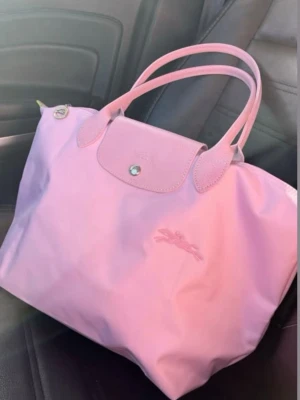 Rosa handväska från Longchamp - Säljer en snygg rosa handväska från Longchamp med klassisk design och silverfärgade detaljer. Väskan har två handtag, lock med knapp och dragkedja. Materialet är en mix av slitstark syntet och skinn på handtag och lock. Perfekt storlek för att få plats med allt du behöver.