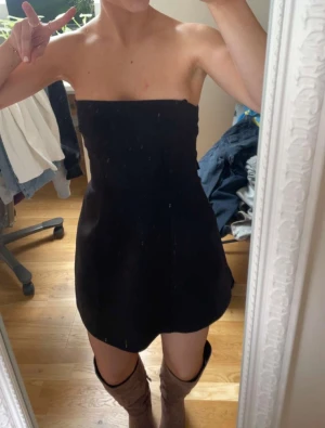 Svart kort klänning med rosett - Supersnygg svart kort klänning från Zara med axelbandslös design och stor rosett i ryggen. Klänningen har en lätt utsvängd form och är tillverkad i ett slätt material som ger en stilren look. Klänningen är hel svart utan fläckar och endast använd 1 gång❤️