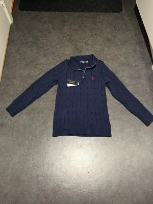 Marinblå stickad tröja Polo Ralph Lauren - Säljer en marinblå stickad tröja från Polo Ralph Lauren med kabelstickat mönster och röd logga på bröstet. Tröjan har lång ärm, ribbade muddar och en half zip-dragkedja vid halsen. Perfekt för dig som gillar klassisk och stilren design. Ett litet hål finns, men verkligen inget som syns när den bärs!