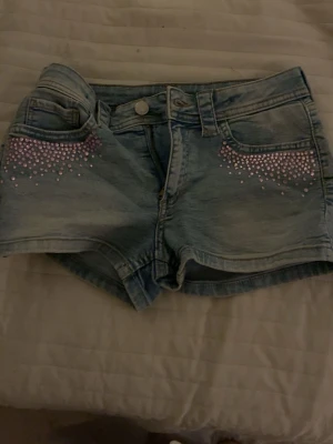 jättesöta jeansshorts H&M - Säljer ett par ljusblå jeansshorts från H&M, storlek 152. Shortsen har en unik design med rosa pärlor och De har knapp och dragkedja framtill. 