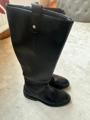 Svarta höga skinnstövlar - Säljer ett par klassiska svarta stövlar i skinn med rund tå och platt sula. Stövlarna har en stilren design med diskreta sömmar och en liten rem upptill med metallknapp. Perfekta till jeans eller kjol för en clean look.