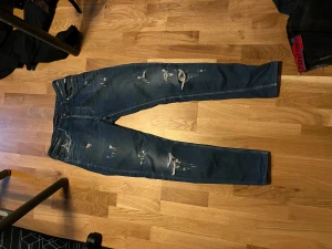 Blå Dsquared2 jeans med slitningar - Säljer ett par blå jeans från Dsquared2 med snygga slitningar och distressed detaljer både fram och bak. Modellen har klassisk femficksdesign, smal passform och låg midja. Materialet är jeans i bomull och de har en cool streetkänsla.